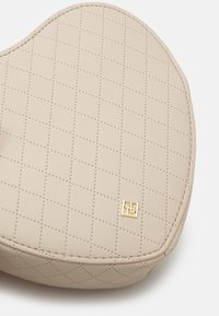 Beżowy pikowany skórzany dodatek z przeszyciami w kształcie diamentów i małym złotym kwadratowym logo na białym tle.