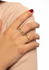 Anillos de oro y plata llevados en una mano, con una banda de oro lisa y una banda de plata con piedras brillantes, sobre un fondo texturizado en color rosa.