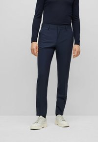 Pantaloni da abito blu navy con una vestibilità su misura, caratterizzati da una texture liscia, frontale piatto, tasche laterali e passanti per cintura, abbinati a scarpe da ginnastica bianche.