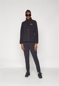 Black fleece jacket with high collar, zip closure, and logo; paired with black pants featuring side zippers, worn with black sneakers.