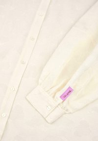 Camicia bianca con bottone e motivo floreale testurizzato, caratterizzata da piccoli bottoni e maniche arricciate, etichettata "OH APRIL" su un cartellino rosa.