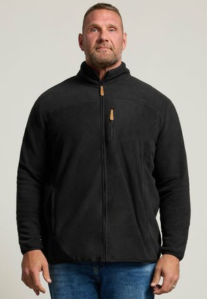 Homme debout face à l'avant, portant une veste polaire noire zippée et un jean bleu sur un fond uni.