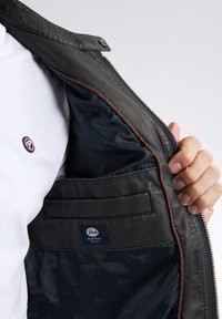 Schwarze Lederjacke mit einem Reißverschluss vorne und braunen Akzenten. Das Innenfutter verfügt über eine Tasche und ein blau gemustertes Futter, mit einem sichtbaren Logosticker.