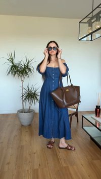 Femme en robe bleue épaules dénudées et sandales ajustant ses lunettes de soleil, tenant un grand sac à main marron, debout sur un sol en bois près d'une plante.