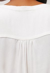 Primo piano di una blusa bianca strutturata sul retro con una spallina e tessuto arricciato sotto il collo su una persona con capelli scuri.