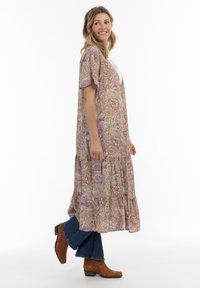 Robe longue à motifs paisley en beige, avec des manches courtes et un ourlet à niveaux. Associée à un jeans noir et des bottines en daim marron.