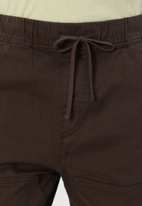 Brown elastic-waist joggers with a drawstring, featuring side pockets and a smooth texture. Close-up view focuses on waist and fabric details.