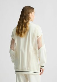 Weißjacke mit durchsichtigen, gemusterte Ärmeln und einem kontrastierenden schwarzen Streifen am Saum. Glattes Material mit lockerer Passform und offenem Rückendesign.