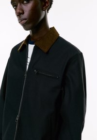 Black jacket with a brown corduroy collar, featuring a zippered front and chest pocket. The material appears textured and durable.