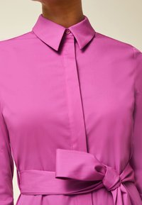 Abito rosa con colletto e chiusura anteriore a bottoni, dotato di una cintura legata in vita e di una texture di tessuto liscia e leggera.