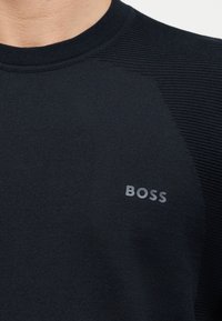 Detailní záběr osoby nosící černý texturovaný pletený svetr s logem "BOSS" na hrudi.