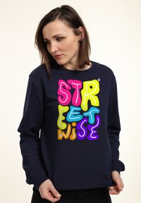 Námořnický svetr s barevným, hravým textovým grafickým prvkem, který zní "STREETWISE" v růžové, žluté, zelené, modré, oranžové a fialové. Dlouhé rukávy.