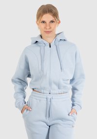 Felpa corta azzurro chiaro con zip frontale, cordino regolabile e polsini a coste. Abbinata a pantaloni della tuta coordinati, entrambi in materiale morbido.