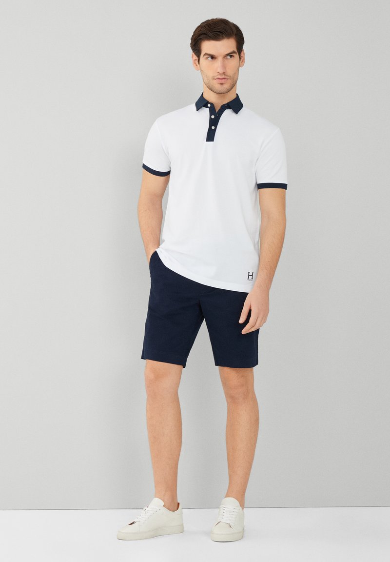 T-shirt polo blanc avec col et poignets marins, associé à un short marine. Le mannequin se tient dans des baskets blanches, affichant une tenue décontractée.