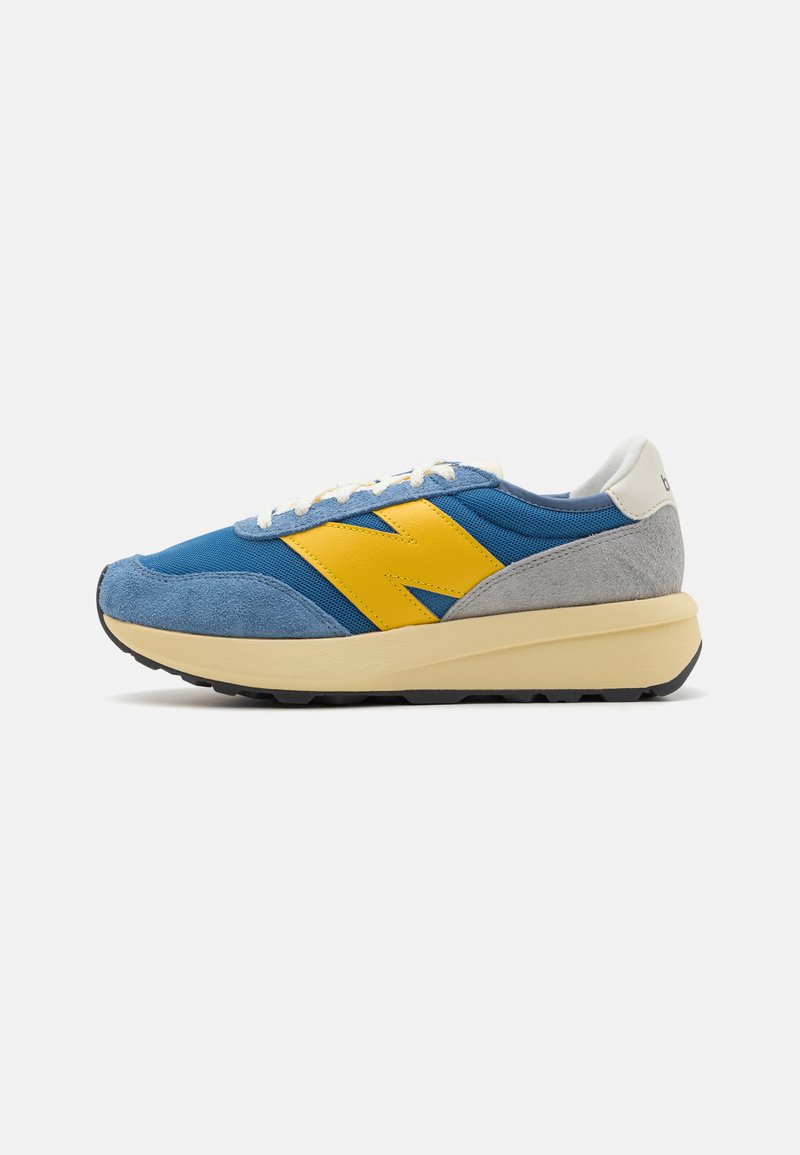 Zapatilla azul y amarilla que presenta un upper de ante y malla, una suela acolchada y un distintivo logo amarillo 'N' en el lateral.