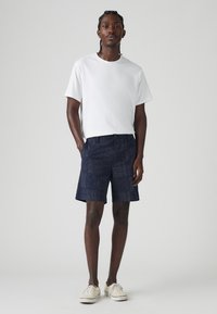 Tricou alb cu mânecă scurtă, asortat cu pantaloni scurți navy cu un design patterned, având o textură interesantă. Încălțăminte sport de culoare deschisă completează ținuta.