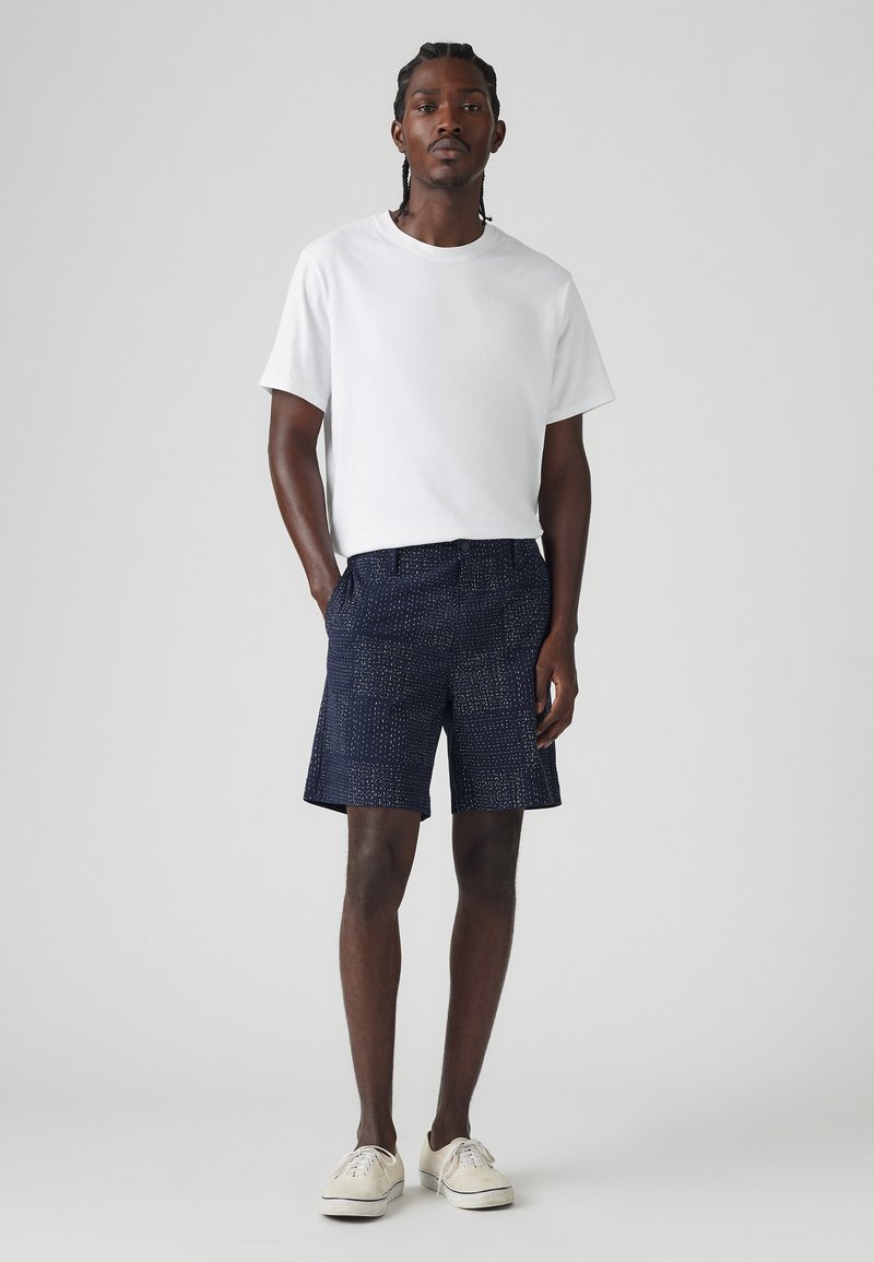 Tricou alb cu mânecă scurtă, asortat cu pantaloni scurți navy cu un design patterned, având o textură interesantă. Încălțăminte sport de culoare deschisă completează ținuta.