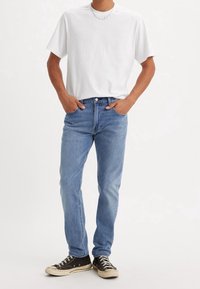 Homme portant un t-shirt blanc uni, un jean bleu clair, des baskets noires et une fine chaîne, debout les mains dans les poches.