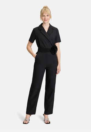 Combinaison noire à manches courtes, avec un col ajusté, une ceinture large ornée d'une accent floral et un pantalon à jambes larges. Portée avec des sandales à lanières.