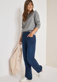 Jeans a zampa di elefante blu navy con un fit classico, abbinati a un maglione grigio chiaro e sneakers bianche. Una giacca in velluto a coste beige è tenuta in una mano.