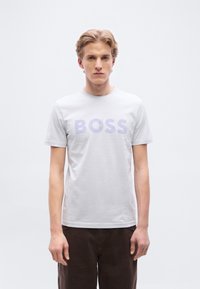 Ανοιχτό γκρι βαμβακερό μπλουζάκι με μεγάλο λογότυπο "BOSS" σε λιλά χρώμα στο στήθος, συνδυασμένο με σκούρο καφέ παντελόνι και ουδέτερο φόντο.