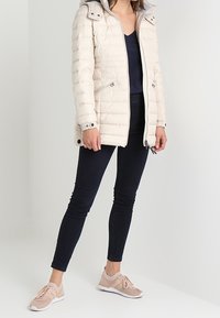 Lys-beige pufferjakke med hætte, lynlås i front og sidelommer. Båret sammen med mørke skinny jeans og lyserøde sneakers med detaljer.