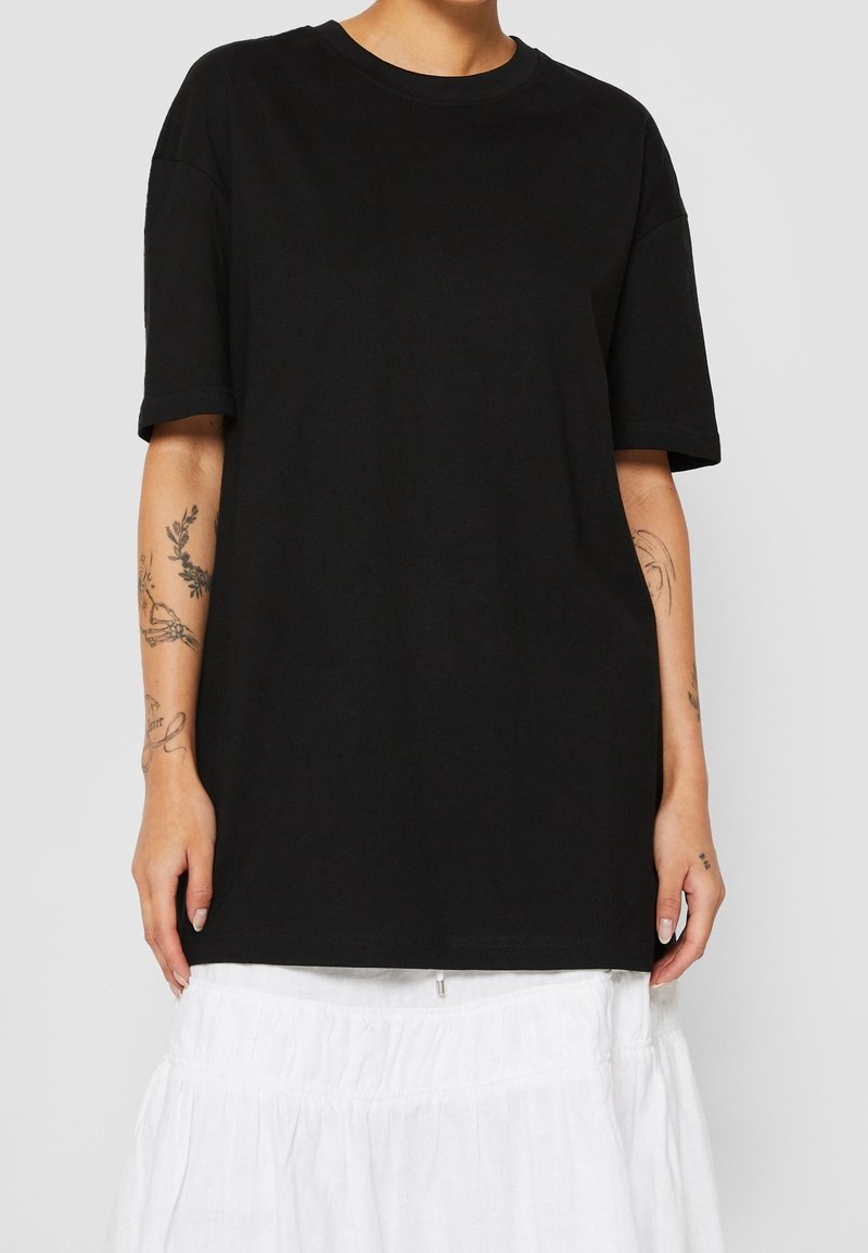 Personne portant un t-shirt noir uni oversize à manches courtes, avant-bras tatoués et jupe blanche sur un fond uni.
