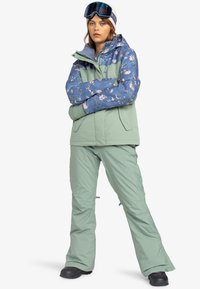 Snowboard jacket with a floral blue and green design, paired with matching green pants. Features zippered pockets and a hood.