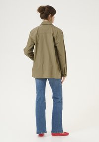 Olive zeleno oversize srajcni plašč z ovratnikom, gumbnimi manšetami in ravnim robom, v kombinaciji z modrimi ravnimi kavbojkami in rdečimi balerinkami.