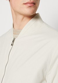 Giacca bomber leggera color crema con chiusura a zip, tessuto testurizzato con un sottile motivo a griglia e colletto a coste.