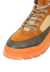 Braune Wildleder- und graue Leder-Schuh mit einer orangen Gummisohle, ausgestattet mit beigefarbenen Mesh-Einsätzen und runden Schnürsenkeln. Dicke, strukturierte Sohle.