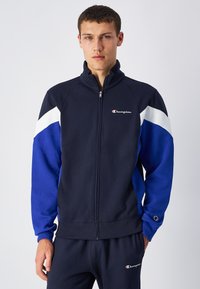 Giacca a zip in tuta di colore navy e blu con dettagli bianchi, colletto alto e un piccolo logo sul petto; tessuto morbido con un design sportivo.