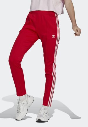 Person trägt rote Adidas Trainingshose mit weißen Streifen und Logo, kombiniert mit weißen Sneakers und Socken, steht vor einem einfarbigen Hintergrund.