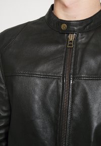Giacca in pelle nera con una texture liscia, dotata di colletto alto, chiusura frontale con zip e dettagli di cucitura sottili. Accenti in ottone.