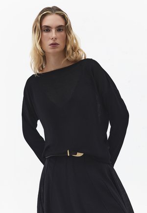 Vrouw met blond golvend haar die een losse zwarte top met lange mouwen en een zwarte hoge taille rok met riem draagt, tegen een witte achtergrond.