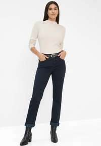 Beige ribbed Langarmshirt, marineblaue schmal geschnittene Hose mit aufgerolltem Saum, schwarze Stiefeletten und ein gemusterter Gürtel mit einer metallischen Schnalle.