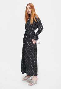Schwarzes Langarmkleid mit einem Gürtel, das ein sich wiederholendes Silber- und lila Muster aufweist. Kombiniert mit silbernen Riemchenheels.