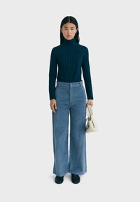 Marineblauwe geribbelde coltrui gecombineerd met lichtblauwe wijde corduroy broek; model houdt een crèmekleurige handtas vast, zwarte schoenen, neutrale achtergrond.