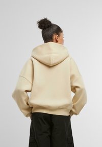 Bež dukserica s kapuljačom, opuštenog kroja, teksturirane tkanine i rebrastih rukava. Model je nosi uz tamne cargo hlače.