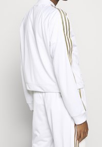 Witte trainingsjack van gladde stof, met drie gouden strepen langs de mouwen en een geribde zoom. Ritstassen aan de zijkanten.