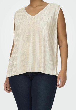Sleeveless cream textured V-neck top worn with dark blue jeans, showing right arm tattoo and relaxed fit from shoulders to hips.