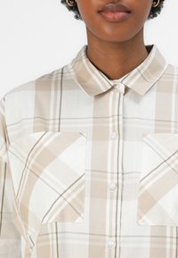 Αεροδιαπερατός καμηλό καρό πουκάμισο με κλασικό γιακά, κουμπιά μπροστά και δύο μεγάλες τσέπες στο στήθος. Απαλή ύφανση με διακριτική γυαλάδα.