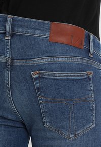 Vista trasera de una persona que lleva jeans de mezclilla azules con un parche de cuero marrón y costuras naranjas en el bolsillo trasero.