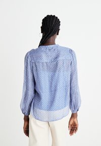 Svetlo modra bluza s geometrijskim vzorcem, dolgimi napihnjenimi rokavi in zaobljenim robom. Narejena je iz prosojnega materiala s sproščenim krojem.