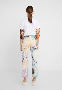 Tie-dye flare jeans met meerkleurige spiraalpatronen; gecombineerd met een wit T-shirt en een kleurrijke lange onderlaag. Witte sneakers.
