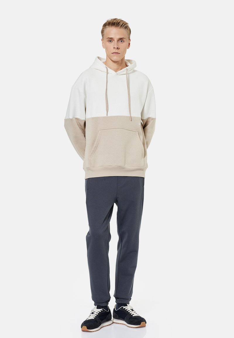 Giovane uomo in piedi con le mani dietro la schiena, che indossa una felpa con cappuccio bianca e beige, pantaloni da jogging scuri e scarpe da ginnastica nere con lacci bianchi.