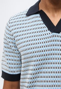 Άνδρας που φοράει πλεκτό μπλουζάκι με κοντά μανίκια, με γεωμετρικό μοτίβο σε μπλε, μαύρο και λευκό, μαύρο γιακά και μαύρα μανσέτες στα μανίκια.