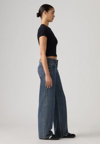 Μαύρο κοντοκομμένο μπλουζάκι συνδυασμένο με φαρδιά μπλε τζιν παντελόνια. Τα τζιν έχουν ψηλή μέση και εμφανείς ραφές, δημιουργώντας μια χαλαρή σιλουέτα.