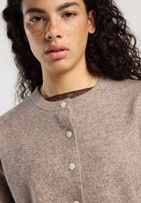 Nuori henkilö, jolla on kihara tumma hiukset ja pisamia, pukeutunut nappikiinnitteiseen vaaleanbeigeen neuleeseen ja ohueen kultaketjuun.