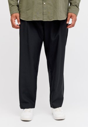 Homme portant un pantalon noir ample, une chemise verte boutonnée et des baskets blanches, debout devant un fond blanc.